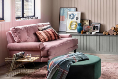 A pink cottage style living room with shiplap panelling, a pink fabric sofa and dark green velvet effect pouffe.