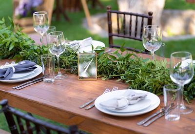 Wooden dining table in a garden decorated with a green garland centrepiece with plates, cutlery, napkins and glasses.