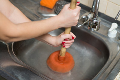 Plunger sat on top of a plughole and partially submerged in water. Person is gripping the plunger with two hands and pushing it down.