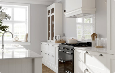 A galley kitchen is finished in soft, creamy white shaker-style cabinets with a matt finish. There are wall-mounted glass-front cabinets, and an island with a white farmhouse sink. The countertops are white and grey marble-effect.