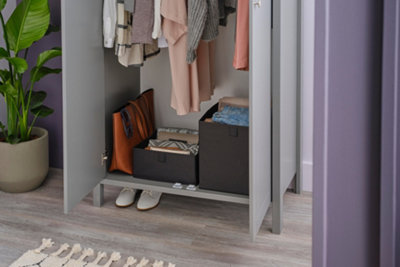 A small purple bedroom with a slim grey wooden wardrobe with open doors, full of clothes and storage boxes.