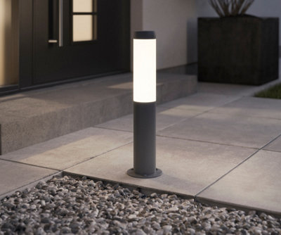 A small anthracite post light installed on a paving slab outside the modern entrance of a home.