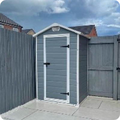 A beige garden shed with dark double doors is placed at the end of a neat backyard featuring artificial grass, potted plants, and modern landscaping elements. The space is framed by fencing and brick homes in the background. This is user-generated content shared from a real garden on Instagram with the handle @millenniummoseley