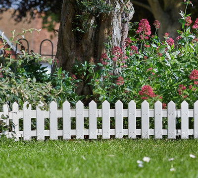 A white picket fence in a garden, separating a border of trees and large shrubs from a lush green lawn.