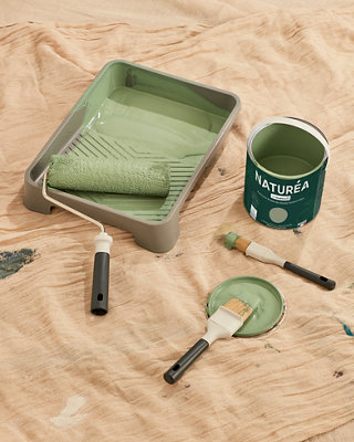 A paint tray with a pot of paint and a paintbrush covered in green paint is placed on top of a brown dust sheet.