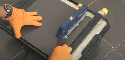 Person wearing gloves feeding a tile into the blade of an electric tile saw to cut it. Water from the saw is visible on top of the tile.
