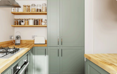 A kitchen is bathed in light from a window. The kitchen is L-shaped and features shaker style cabinets in a pale green colour with a matt finish. At the end of the unit there are two tall cabinets. The countertops and open shelves on the wall have a warm wood finish.