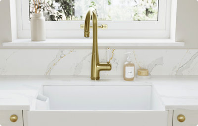 A white kitchen sink pairs with a white marble-effect countertop and splashback. A brass kitchen tap glistens in the sunshine from the window behind.