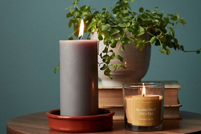 A large grey pillar candle and small yellow scented candle in a glass jar on a wooden side table.