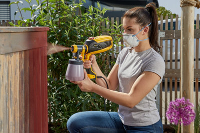 Person wearing a dust mask using a paint sprayer to paint a section of garden fence.