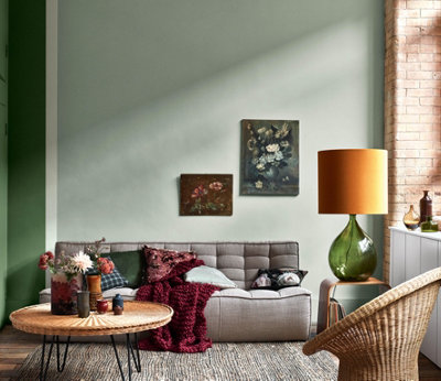 A green living room with a grey sofa, a rattan statement chair, round rattan coffee table, deep burgundy soft furnishings and floral wall art.