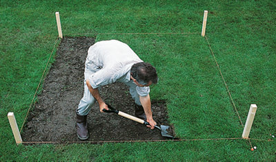 Person using a shovel to remove the grass from an area of ground which has been marked out by 4 wood pegs and a builders line.