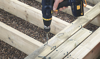 Person using a drill to drive a decking screw through a decking board to secure it to the decking joist below. Other boards have already been installed around the board that is being worked on.