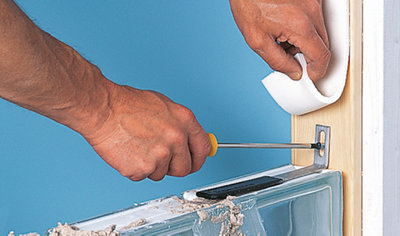 Person using a screwdriver to secure a panel anchor to the timber frame surrounding a glass block wall. Panel anchor sits on top of the first glass block of the row.