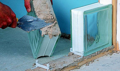 Using a trowel to lay mortar onto the bottom of a glass block. The first glass block in the row has already been installed and the spacer for the second block is already in the mortar.
