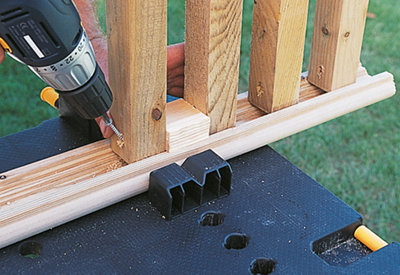 Using a drill to drive a screw diagonally through a spindle into the underside of a handrail. The spindles are spaced apart using a piece of timber as a spacer.