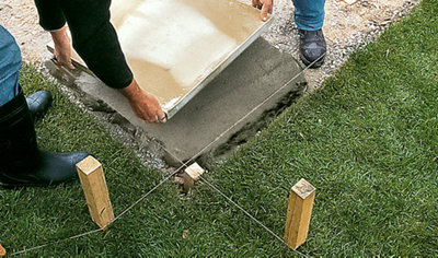 Two people lifting a paving slab and carefully placing it onto the mortar in the corner of an area that has been marked out for a shed base.
