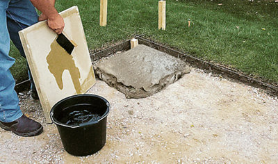 A person using a brush to wet the back of a paving slab, preparing it for laying on top of the mortar that has already been spread on the ground.