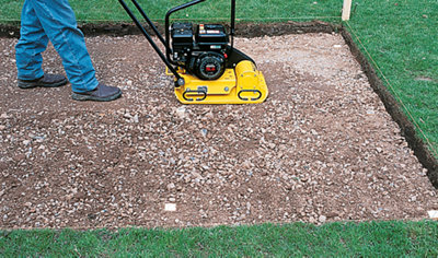 A person using an earth rammer to compact the hardcore in a prepared area, ensuring that it is level.