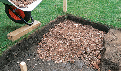 A person using a wheelbarrow to tip hardcore into an excavated area, which is being prepared as the sub-base for a shed base.