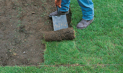 A person using a shovel to remove turf from an area that has been marked out with a builder's line and pegs.