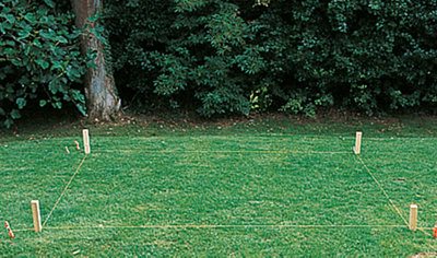 A square area on a lawn marked out with wooden pegs and a builder's line, indicating the planned location for a shed base.