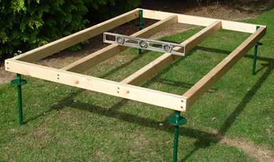 A wooden shed base made up of six pieces of timber arranged in a grid pattern, with four metal spikes at each corner for securing it into the ground.