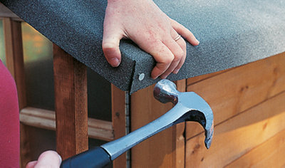 A person using a hammer to drive nails through the roofing felt into the wooden playhouse.