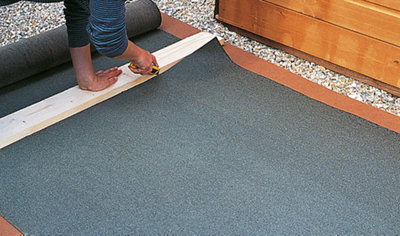 A piece of timber is laid on top of roof felt, which is placed on the floor. A person is using a utility knife to cut the felt along the edge of the timber.