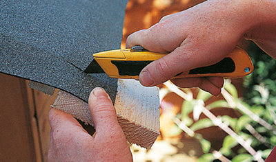 A person is holding a piece of timber underneath the overhang of roofing felt while using a utility knife to cut the felt.