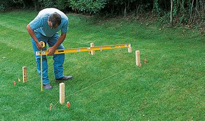 A person resting a spirit level on a peg at the top of a slope. The spirit level is held level while they measure the distance from the spirit level to the ground at the bottom of the slope.