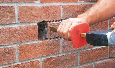 Using a chisel and club hammer to remove a half brick from a wall where holes have been drilled.