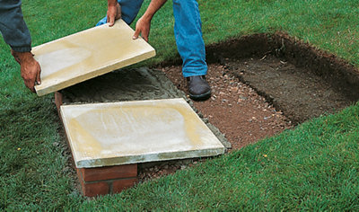 Two people lowering a paving slab onto a bed of mortar, laid on top of a hardcore base and a course of bricks. The slab is positioned next to another slab to form the bottom step.