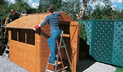 Two people stood on step ladders positioning a shed roof panel onto the top of a shed.