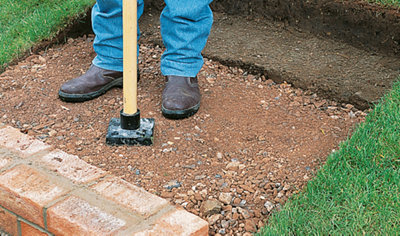 A person using an earth rammer to compact hardcore laid behind a course of bricks.
