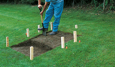 A person using a shovel to shape the outline of garden steps in an area marked with a builder's line and pegs.