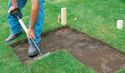 A person using a shovel to remove grass from an area marked with a builder's line and pegs.