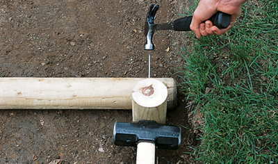 Using a hammer to knock nails through a log stair tread into the supporting stake, which is held in place by a sledgehammer.