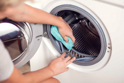 Pulling back the grey seal of a washing machine to clean out dirt that is trapped in the fold using a blue cloth.