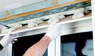 A person placing a piece of plasterboard with adhesive on the bottom onto the reveal above a window.