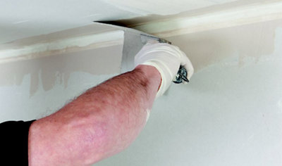 A person using an internal corner trowel to smooth jointing compound applied on top of an angle bead installed between a wall and ceiling.