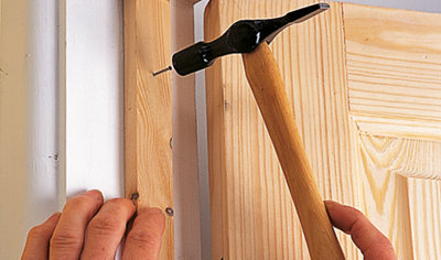 A person using a hammer to drive a nail, fastening a doorstop to a door frame.