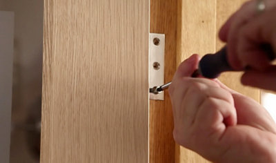 A person using a screwdriver to drive a screw through the hole in a door hinge, securing it to the wall.