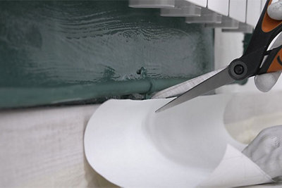 Cutting a piece of wallpaper with scissors to fit underneath a pipe that runs along the wall.
