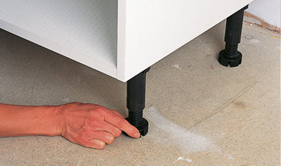 A person adjusting the height of a kitchen cabinet by rotating the adjustable leg at the bottom while ensuring it aligns with the guidelines marked on the wall.