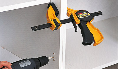 A person using a drill to drive a screw through the side of one cabinet into another to secure them together. The two cabinets are clamped tightly to ensure proper alignment.