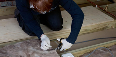 Person wearing gloves and a dust mask using a pair of scissors to cut loft insulation to size around a joist.