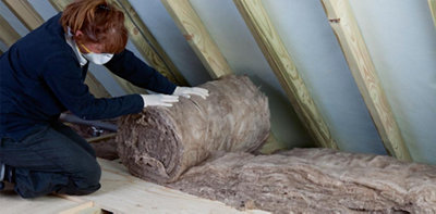Person wearing gloves and a dust mask unrolling a loft insulation roll on top of loft insulation which has already been installed between the joists.