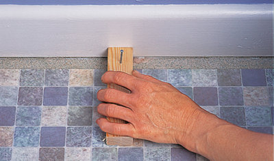 Block of wood with a nail hammered in it pushed up against skirting board on top of vinyl sheet.