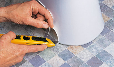Using a utility knife to trim a piece of excess vinyl floor around the base of a toilet.